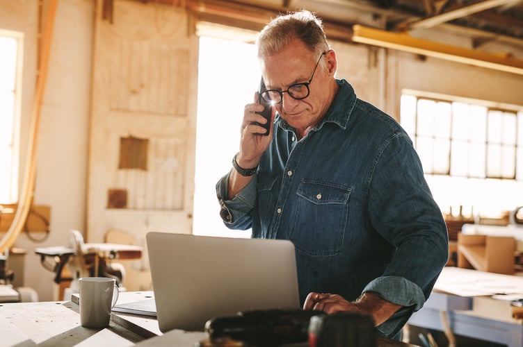 Senior man talking on cell phone and using laptop on work table. Mature carpenter working on laptop and answering phone call in his carpentry workshop.